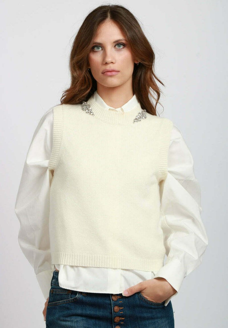 Gilet lavorato a maglia color crema con design senza maniche, caratterizzato da un colletto a coste e dei dettagli decorativi in argento sulle spalle. Camicia bianca sotto.