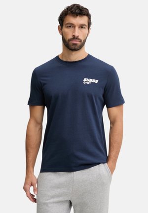 Uomo con capelli corti e ricci e barba, indossa una t-shirt Guess 1981 blu navy e pantaloni della tuta grigio chiaro, in piedi con una mano in tasca.