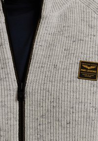 Pleteni sivi dukserica sa zatvaračem, koja ima teksturirani uzorak. Sadrži tamni zatvarač i crno-zlatnu oznaku s logotipom "PME LEGEND" na prsima.
