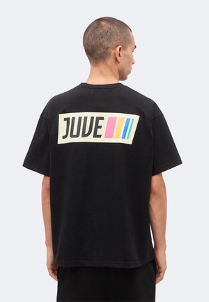 Moški s kratkimi lasmi, ki nosi črno ohlapno majico z napisom "JUVE" in tremi pisanimi navpičnimi črtami na hrbtu.