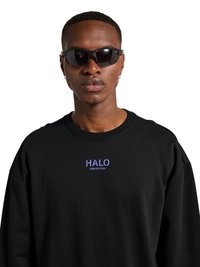 Mężczyzna w czarnych okularach przeciwsłonecznych, złotych kolczykach kołach i czarnej bluzie z fioletowym napisem "HALO" na białym tle.