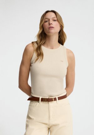 Femme aux longs cheveux blonds, portant un haut sans manches côtelé crème, un pantalon crème et une ceinture en cuir marron sur un fond clair uni.