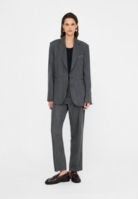 Completo a due pezzi in tessuto grigio scuro a righe, caratterizzato da un blazer aderente con rever a disegno e pantaloni dritti, abbinato a scarpe scure.