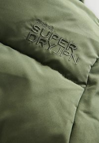 Grøn polstret jakke med en glat tekstur og broderet "SUPER DRY JPN" logo. Letvægtsmateriale og quiltet design til isolering.