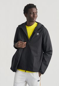 Νέος άντρας που φοράει μαύρο μπουφάν Adidas, κίτρινο πουκάμισο, άσπρο παντελόνι Adidas και κουβαλάει μια μαύρη τσάντα Adidas στον ώμο του.