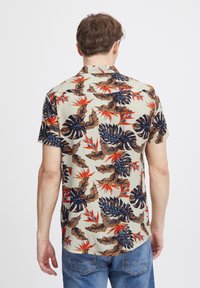 Camisa de manga corta con un fondo beige y un llamativo patrón de hojas tropicales en tonos marino, marrón y rojo; diseño de cuello estándar.
