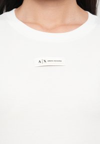Valkoinen puuvillatee paita, jossa on pyöreä kaula-aukkosuunnittelu ja ommeltu etiketti, jossa on mustalla tekstillä "A|X Armani Exchange" -brändäys.