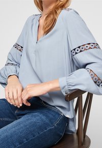 Vaaleansininen pusero, jossa on V-pääntie, puffihihat ja pitsiyksityiskohtia. Pehmeä kangas ja rento malli, yhdistetty tummaansinisiin farkkuihin.