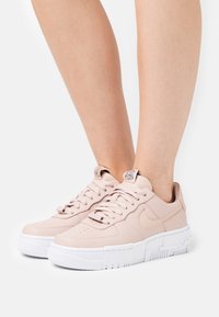 Světle růžové tenisky Nike s bílými tlustými podrážkami, nošené na bosých nohou, zobrazené zepředu a ze strany.