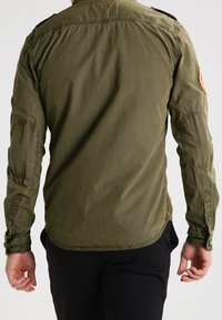 Camicetta utilitaria verde oliva con maniche lunghe, cuciture visibili, una toppa sulla spalla sinistra e orlo curvo. Il tessuto sembra leggero e strutturato.