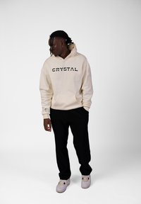 Felpa color crema con testo "CRYSTAL" in nero. Presenta una tasca frontale e cappuccio, abbinata a pantaloni neri a coste e sneaker chiare.