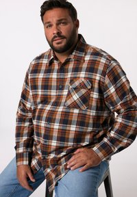 Camicia a quadri marrone e blu navy con colletto bottone, maniche lunghe, tasca sul petto e una texture morbida, abbinata a jeans azzurri chiari.