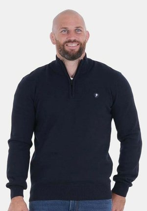 Homme chauve souriant avec une barbe, portant un pull bleu marine à fermeture éclair de quart et un jean bleu, debout devant un fond clair uni.