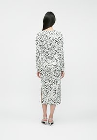 Robe ajustée à manches longues en blanc avec des éclats noirs. Elle présente des fentes sur les côtés et une texture lisse, se terminant au milieu du mollet.