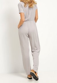 Szary kombinezon z krótkim rękawem, elastycznym pasem, bocznymi kieszeniami, gładką teksturą i prostymi nogawkami, w połączeniu z jasnobrązowym obuwiem.