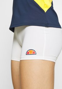 Białe szorty sportowe o dopasowanym kroju, z pomarańczową i niebieską naszywką z logo. W połączeniu z ciemnoniebieską i żółtą górą.