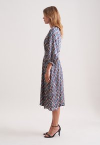 Gemustertes Kleid mit taillierter Oberpartie, ausgestelltem Rock, Dreiviertelärmeln und einer Mischung aus Blau, Rot und Cremefarben. Tragen mit schwarzen High Heels Sandalen.