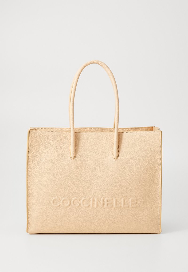 Bež kožna torba s glatkom teksturom, s dva ručka i utisnutim logotipom "Coccinelle" sprijeda. Pravilnog je pravokutnog oblika.