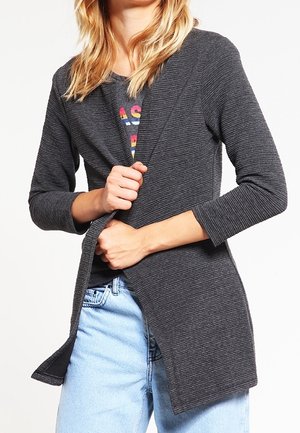 Personne portant un cardigan côtelé gris foncé sur un T-shirt graphique et un jean bleu clair, ajustant le cardigan avec les deux mains.