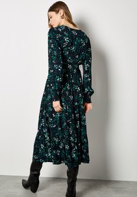 Schwarzes langärmliges Kleid mit grün-weißem Blattmuster, geschichteter Rock und taillierten Bund. Kombiniert mit knielangen schwarzen Stiefeln.