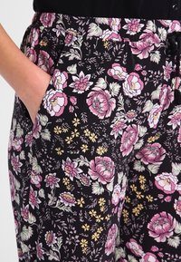 Main dans la poche d'un pantalon noir avec un imprimé floral rose, blanc et jaune, et une taille avec cordon de serrage noir, porté avec un top noir.