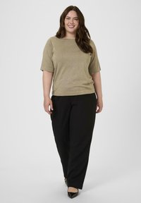 Mujer sonriente con cabello largo y castaño, vestida con una blusa corta de manga beige, pantalones negros y zapatos de tacón negro, de pie contra un fondo sencillo.