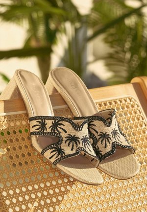 Kreminės spalvos pleišto formos sandalai su juodos palmės siuvinėjimu, padėti ant pintos vytelių kėdės atlošo natūraliame apšvietime.