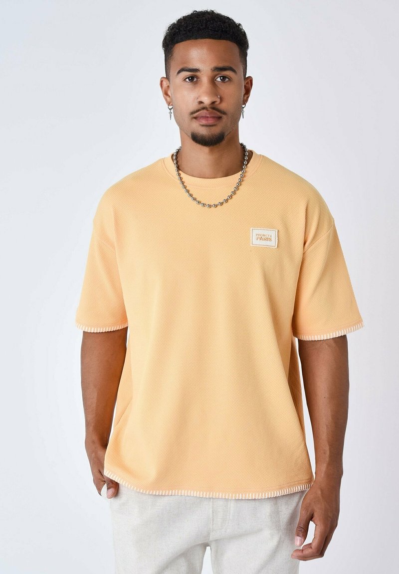 T-shirt a maniche corte, con texture a coste, di colore pesca chiaro, con un logo a patch bianco sul petto e cuciture a contrasto sulle maniche.