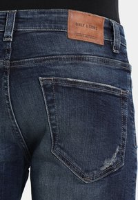 Un jean en denim bleu foncé présente une étiquette en cuir visible portant les mots "ONLY & SONS" sur la taille et une poche arrière avec un léger vieillissement.