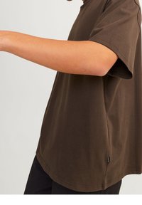 Brun bomull T-shirt med korta ärmar, avslappnad passform, sidoflikar och en liten etikett vid fållen. Slät textur och enkel design.