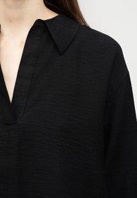 Μαύρο μακρυμάνικο πουκάμισο με υφή, βαθύ V-λαιμόκοψη και μυτερό γιακά, φορεμένο από ένα άτομο με μακριά καστανά μαλλιά.