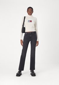 Maglione a collo alto a coste crema con dettagli del logo, abbinato a jeans neri a vita alta. Il modello indossa scarpe platform nere chunky e una borsa nera.