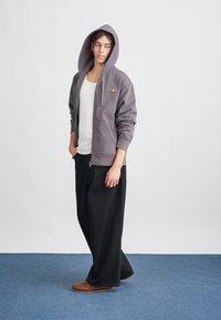 Grå zip-up hoodie med en framficka, bärs över en vit linne. Löst sittande svarta byxor och bruna skor fulländar outfiten.