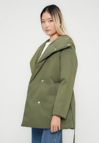 Ung kvinna med långt blont hår som bär en överdimensionerad olivgrön kappa och blå jeans, stående mot en vit bakgrund.