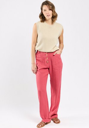 Femme debout portant un haut beige sans manches, un pantalon large rose taille haute avec boutons, et des sandales marron sur fond blanc.