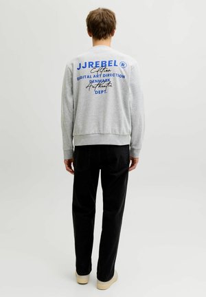 Jeune homme de dos portant un sweat-shirt gris clair avec un texte bleu et noir, pantalon noir et chaussures beiges sur un fond uni.