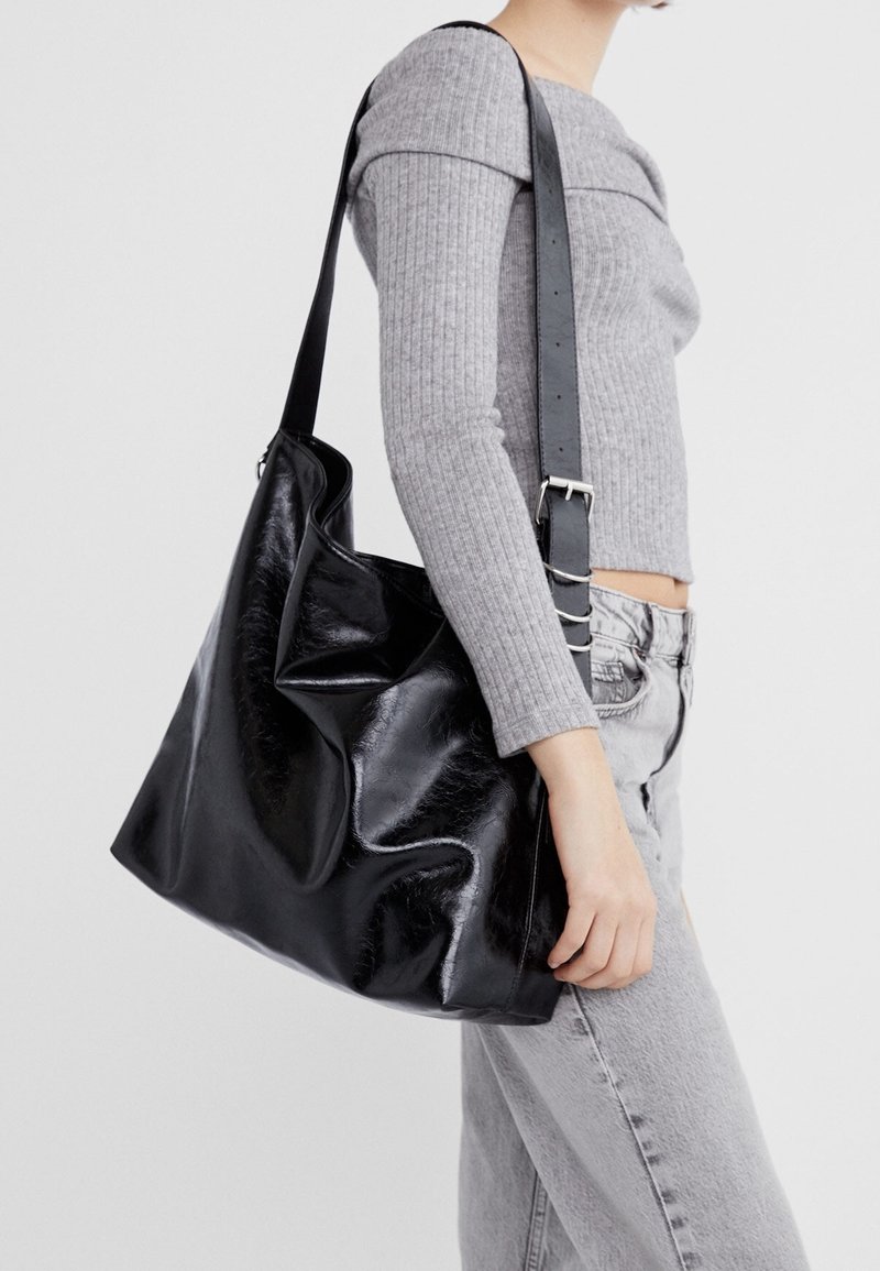 Personne portant un haut gris clair en maille côtelée à manches longues et un jean, porte un grand sac à épaule en cuir noir brillant avec une large bandoulière réglable.