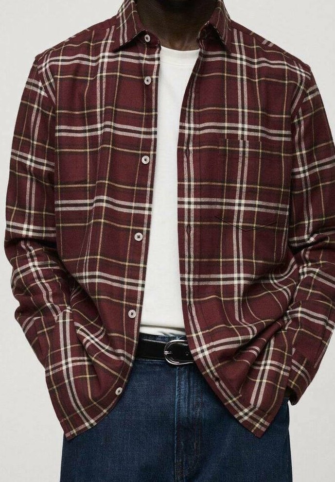 Homme portant une chemise à carreaux bordeaux boutonnée sur un t-shirt blanc, les mains dans les poches, avec un jean bleu et une ceinture noire.