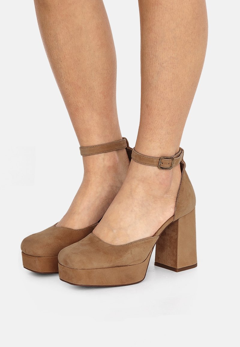 Jambes portant des talons compensés en daim beige avec des brides à la cheville et des talons blocs épais sur fond blanc.