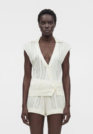 Μαύρη γυναίκα φοράει κρεμ πλεκτό τοπ χωρίς μανίκια με βαθύ V λαιμόκοψη και αντίστοιχα πλεκτά σορτς, στέκεται μπροστά σε απλό λευκό φόντο.