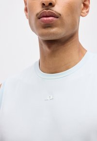 Κοντινό πλάνο ενός ατόμου που φοράει ανοιχτό μπλε αμάνικο πουκάμισο με μικρό λευκό λογότυπο στο στήθος, με φόντο μια απλή επιφάνεια.