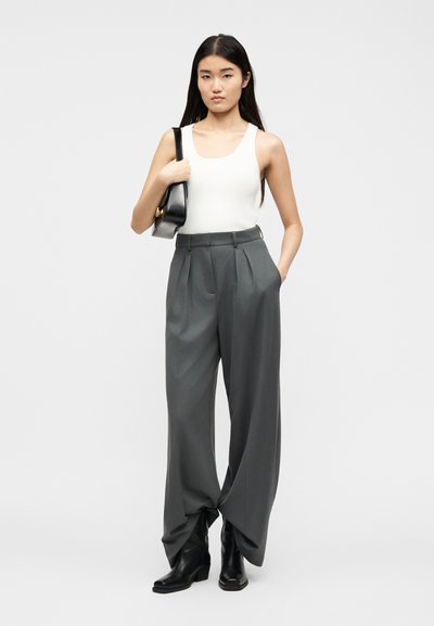 Νεαρή γυναίκα με λευκό αμάνικο τοπ, γκρι φαρδιά παντελόνια με ψηλή μέση, μαύρες μπότες, κρατώντας μαύρη τσάντα ώμου με το ένα χέρι στην τσέπη.