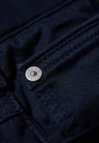 Tessuto blu scuro con una trama liscia, caratterizzato da un bottone in metallo argento con testo inciso e cuciture a contrasto lungo le cuciture.