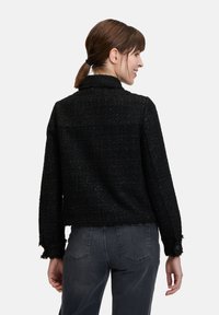 Czarna teksturowana kurtka o skróconym kroju, z kołnierzem, zapinanymi na guziki mankietami oraz postrzępionym dołem. Noszona z ciemnoszarymi dżinsami.