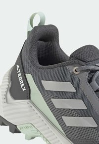 Pantofii sport gri, cu un exterior din plasă texturată, accente verzi, trei dungi albe și o talpă rezistentă, concepută pentru aderență.