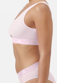 Ljusrosa sport-bh med breda axelband, låg halsringning och ett elastiskt band med en vit Adidas-logotyp. Mjuk, slät textur.