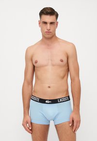 Svetlo modri bombažni bokserci z črnim pasom, na katerem je napis "LACOSTE" in logotip. Oblikovanje je prilegajoče in gladko na otip.