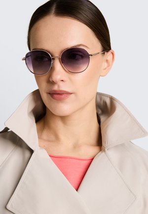 Frau mit glatten dunklen Haaren trägt runde verlaufende Sonnenbrille, korallenfarbenes Oberteil und beigen Mantel mit hochgestelltem Kragen vor einfachem Hintergrund.