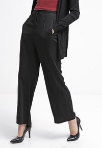 Pantalon noir à jambes larges avec une texture lisse, présentant des plis à l'avant, des poches latérales et une taille haute. Associé à des talons noirs.