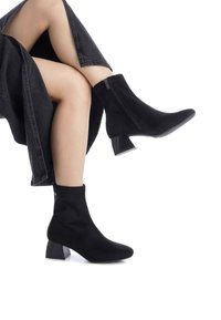 Botas de tobillo negras de suave ante, con punta afilada, cierre de cremallera y un tacón geométrico robusto. Acentuadas con un pequeño detalle de logo.